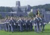 Cadets in gray uniforms march in formation on a campus lawn