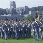 Cadets in gray uniforms march in formation on a campus lawn