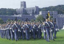 Cadets in gray uniforms march in formation on a campus lawn